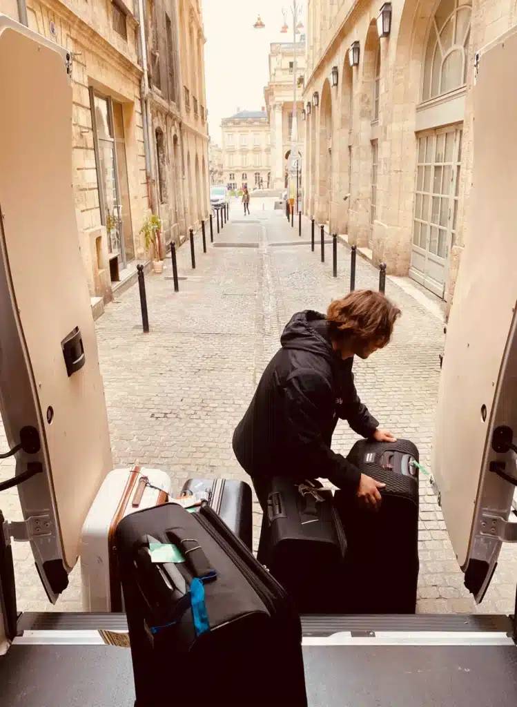 Une personne charge plusieurs grandes valises à l'arrière d'une camionnette dans une rue pavée bordée de bâtiments en pierre et de bornes noires. On aperçoit quelques personnes qui marchent au loin.