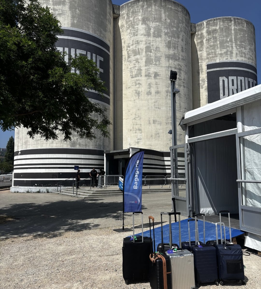 Plusieurs valises sont alignées à l'extérieur d'une tente, près de grands silos cylindriques ornés de bandes noires et blanches, mettant en évidence le stockage de bagages sécurisés. Un drapeau bleu se dresse à proximité, tandis que quelques personnes se rassemblent près de l'entrée du silo, à l'arrière-plan.