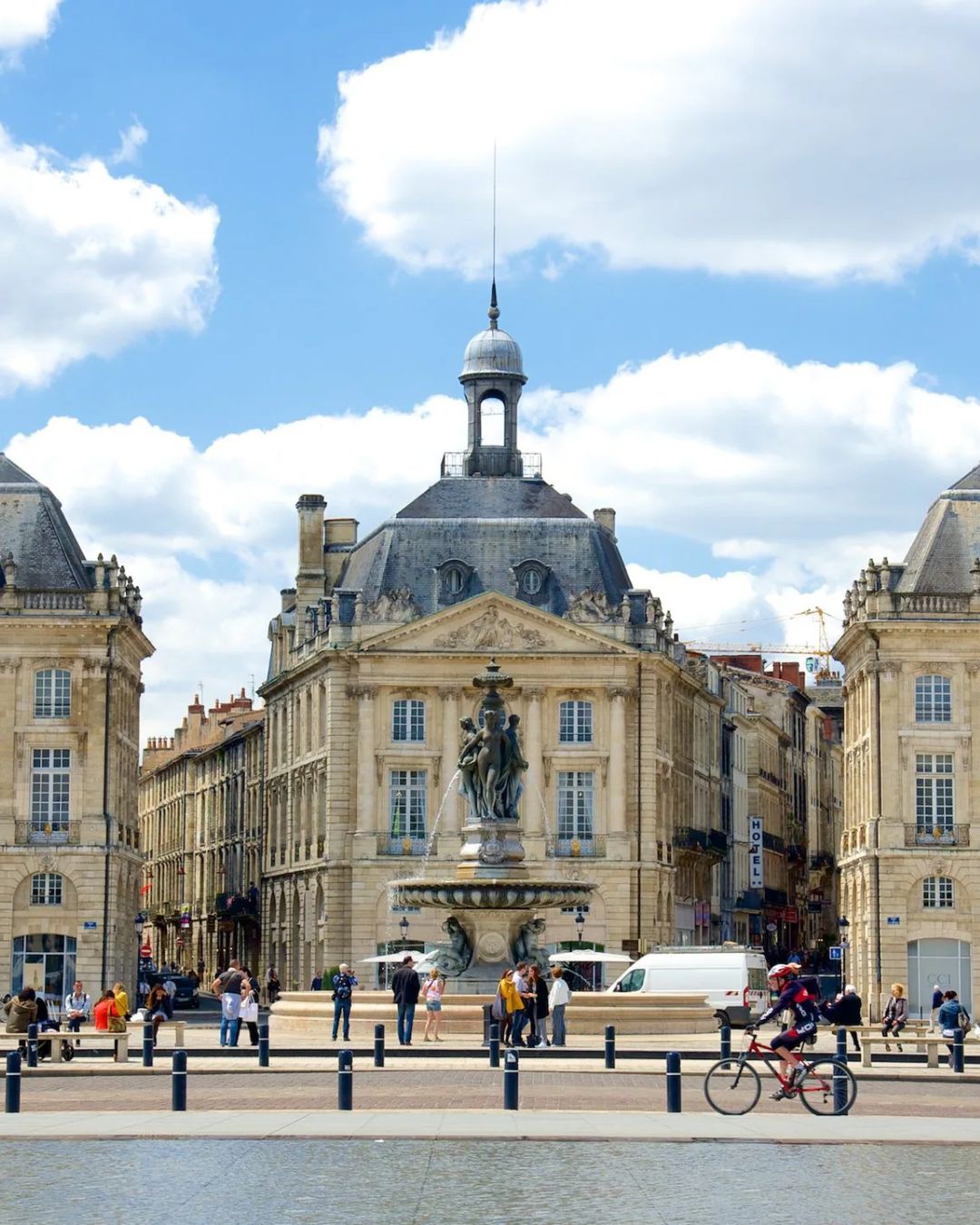 Un grand bâtiment historique avec un dôme se dresse derrière une fontaine et une place, où les gens se promènent et font du vélo sous un ciel bleu. La scène se reflète dans un bassin peu profond, capturant l'atmosphère animée idéale pour le portage bagages événements ou la consigne bagages France services.