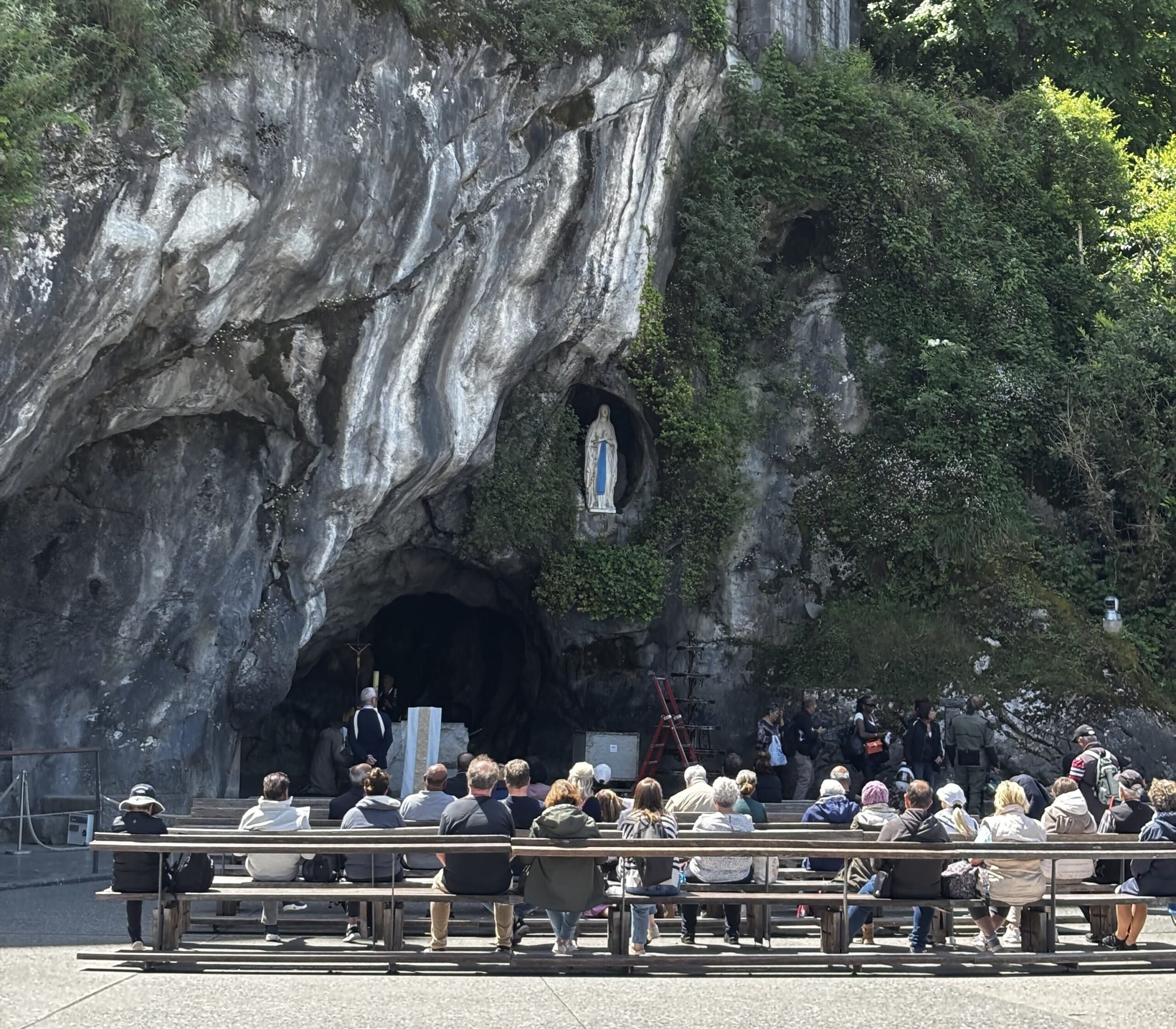 Un groupe de personnes est assis sur des bancs face à une grotte avec une statue de la Vierge Marie encastrée dans la paroi rocheuse, entourée de verdure. Des individus se tiennent à proximité, peut-être en train d'organiser des portages événements pour les visiteurs.