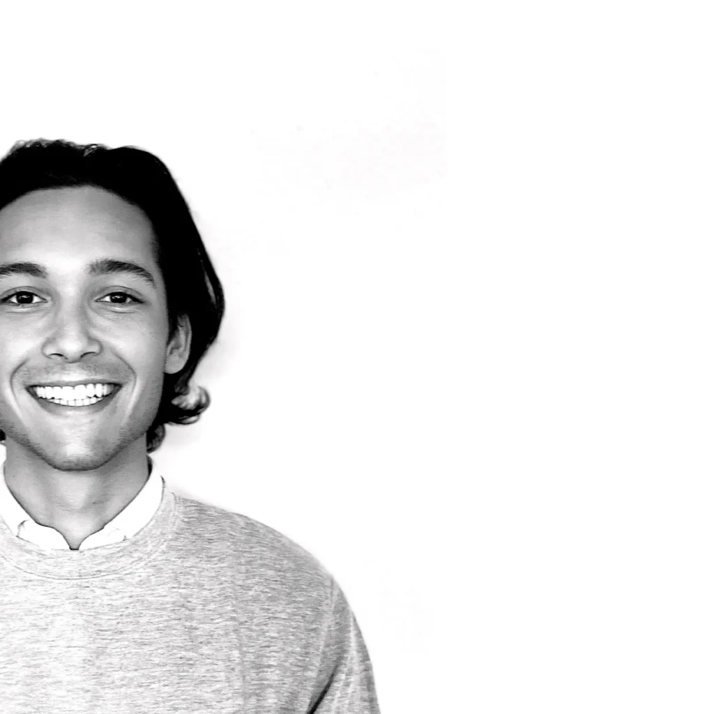 Un jeune homme aux cheveux mi-longs sourit à la caméra. Il porte un pull léger sur une chemise à col. La photo en noir et blanc, sur fond blanc, indique qu'il peut utiliser stockage bagages sécurisé ou consigne bagages France pour des raisons de commodité.
