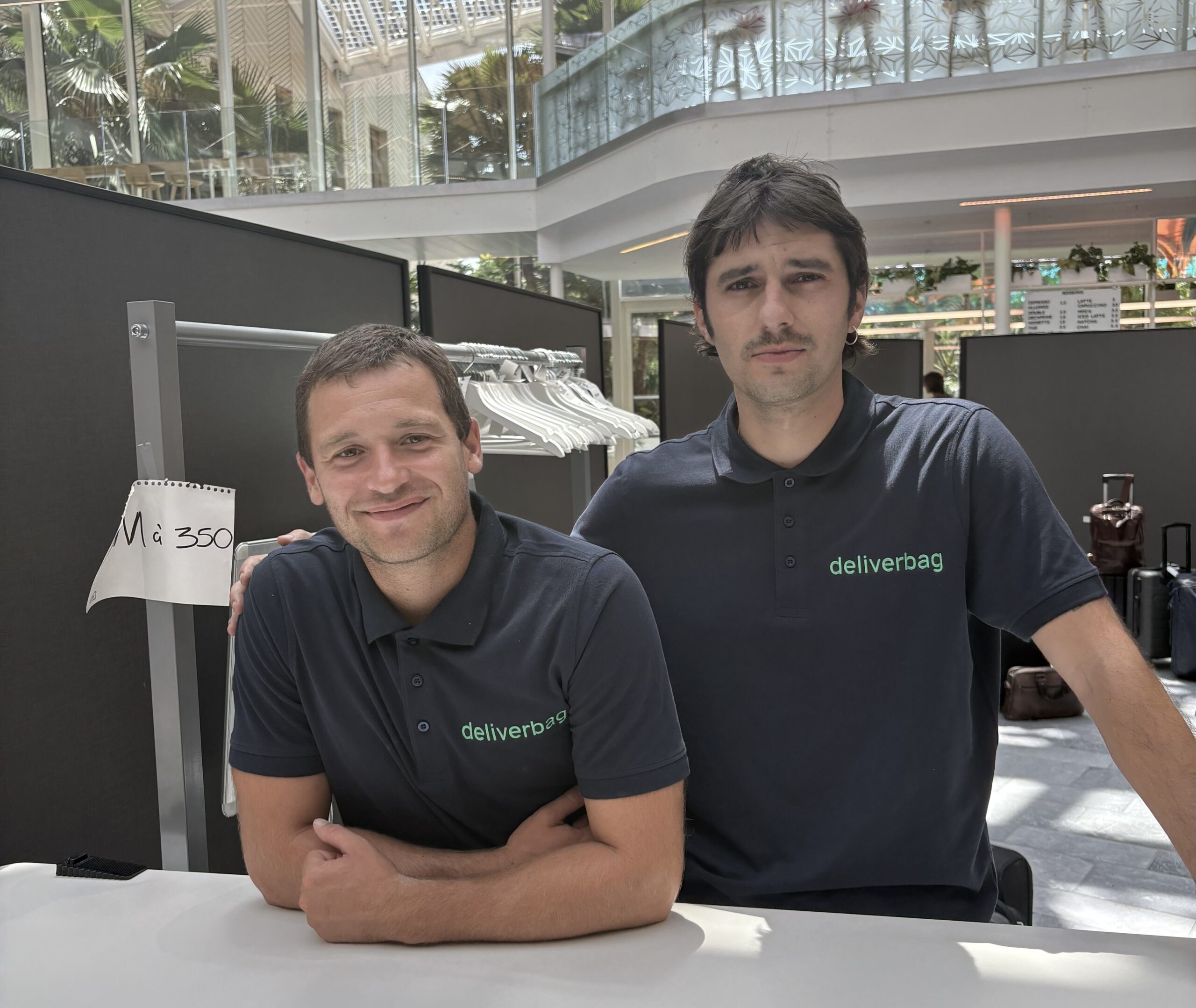 Deux hommes portant des polos bleu marine "deliverbag" sourient et posent à un comptoir dans un espace intérieur lumineux, représentant un service de consigne bagages France, avec des cintres et un panneau manuscrit "Na 350" visible derrière eux.