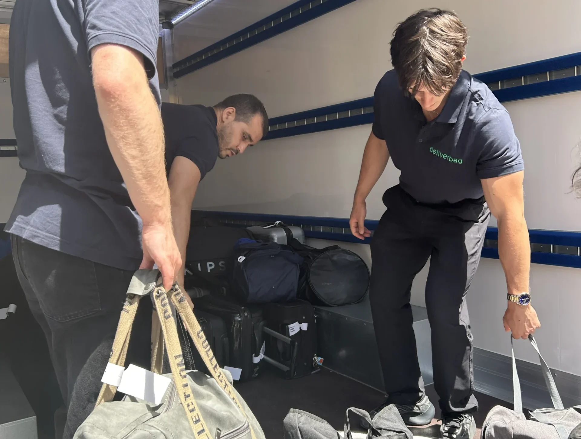 Trois hommes déchargent des sacs de voyage d'un camion dans le cadre d'un service de portage bagages événements. Vêtus de pantalons et de chemises sombres, ils travaillent entourés de sacs à l'intérieur du camion ensoleillé, assurant un transport efficace des bagages Europe.
