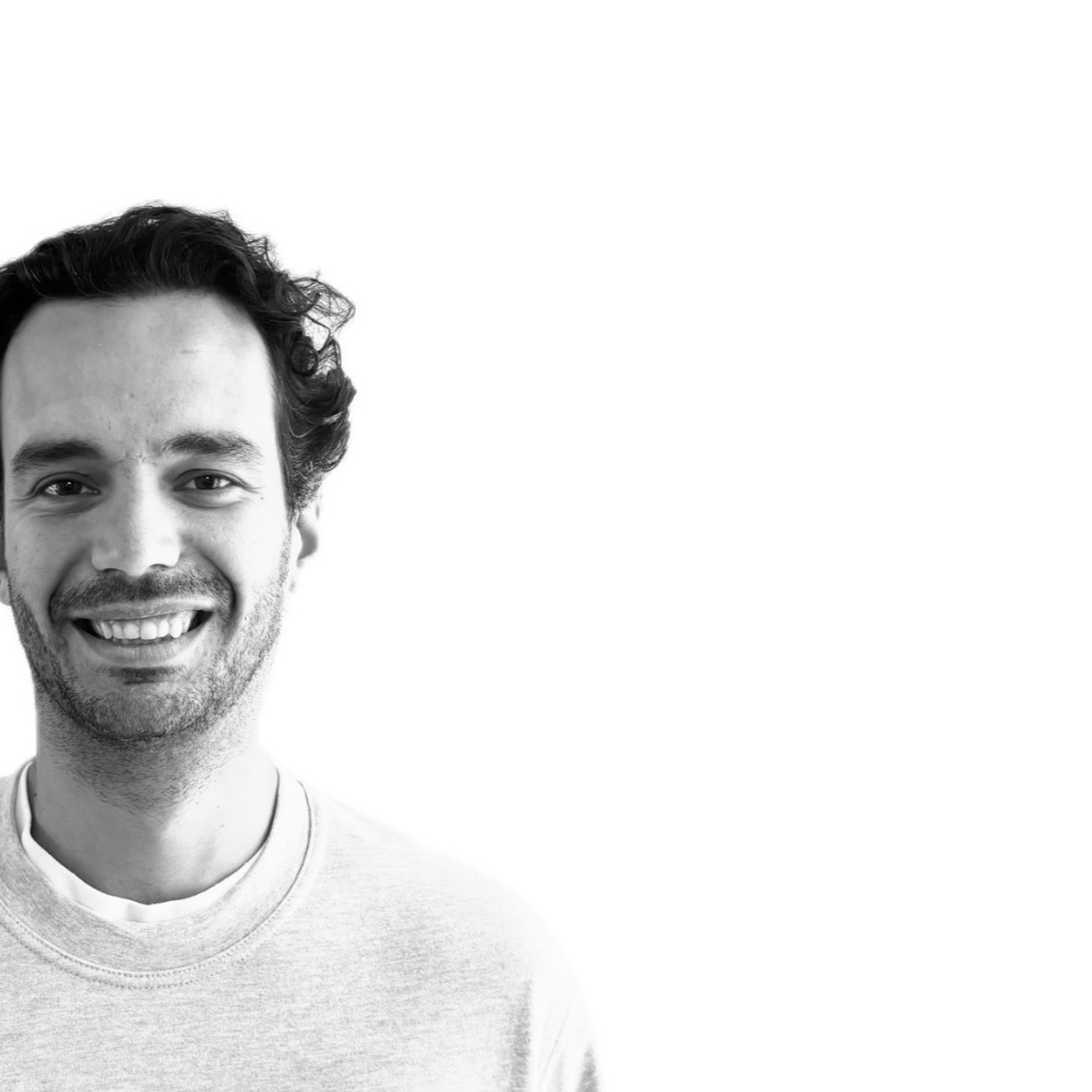Un homme aux cheveux sombres et bouclés et à la barbe sourit. Il porte un sweat-shirt clair sur une chemise blanche. La photo en noir et blanc, sur un fond uni, met subtilement en valeur le portage bagages événements dans sa composition moderne.