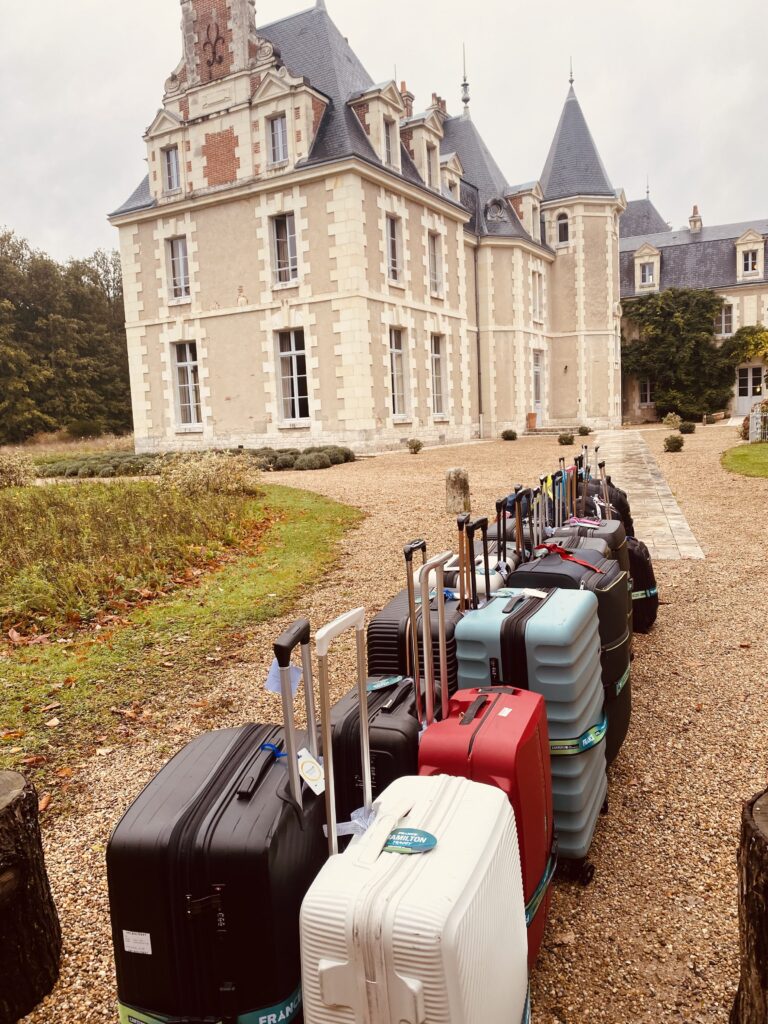 Une rangée de valises colorées alignées sur un chemin de gravier mène à un grand et élégant manoir en pierre avec des toits et des tours en ardoise, entouré d'arbres et de jardins - parfait pour la consigne bagages France. Une rangée de valises colorées alignées sur un chemin de gravier mène à un grand et élégant manoir en pierre avec des toits et des tours en ardoise, entouré d'arbres et de jardins - parfait pour la consigne bagages France.