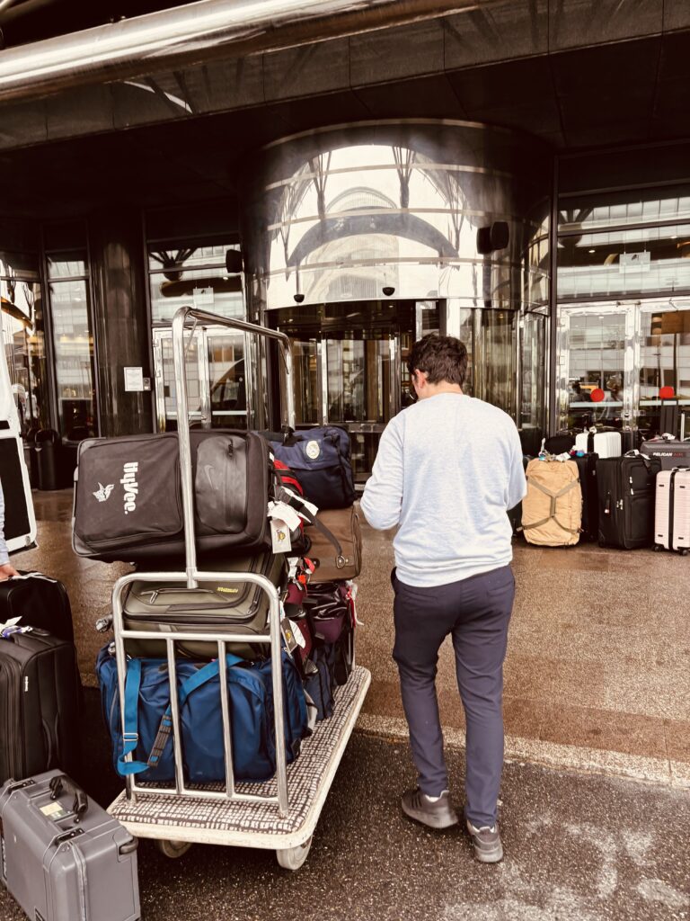 Une personne se tient à côté d'un chariot à bagages empilé devant l'entrée d'un bâtiment avec des portes vitrées, mettant en évidence le portage bagages événements. D'autres valises sont visibles sur le sol à proximité. Une personne se tient à côté d'un chariot à bagages empilé devant l'entrée d'un bâtiment avec des portes vitrées, mettant en évidence le portage bagages événements. D'autres valises sont visibles sur le sol à proximité.