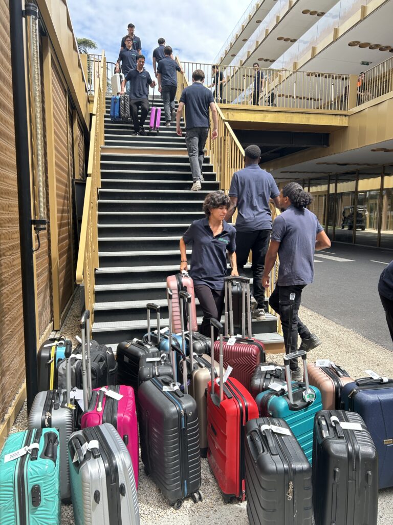 Un groupe en vêtements sombres monte et descend des escaliers extérieurs à côté d'une pile de valises colorées avec des poignées allongées à la base, près d'un bâtiment moderne, illustrant les services de portage bagages événements et de consigne bagages France.
