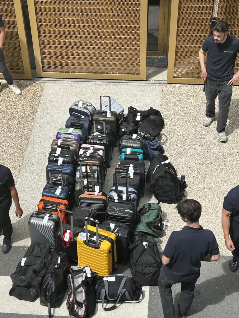 Un groupe de personnes vêtues de chemises sombres se tient autour de plusieurs rangées de valises colorées et de sacs de sport noirs, soigneusement rangés à l'extérieur d'un bâtiment aux portes métalliques, présentant un service de consigne bagages France et de stockage bagages sécurisé.