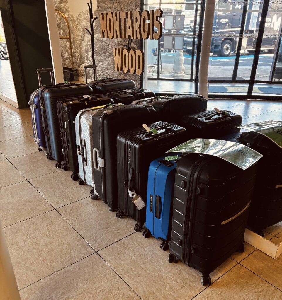 Un groupe de valises de tailles diverses se tient en deux rangées bien ordonnées près d'une fenêtre dans un hall d'entrée. Des panneaux sur le mur indiquent "MONTARGIS WOOD", et un bus est visible à l'extérieur - idéal pour le transport de bagages en Europe et le service de bagagerie mobile.