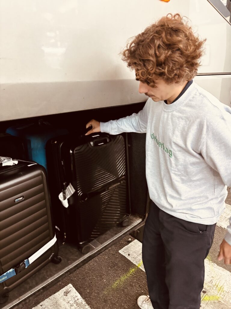 Une personne aux cheveux bouclés, vêtue d'un sweat-shirt gris et d'un pantalon sombre, charge une valise noire dans le compartiment de rangement d'un autobus - montrant une bagagerie mobile de service avec plusieurs autres sacs visibles à l'intérieur.