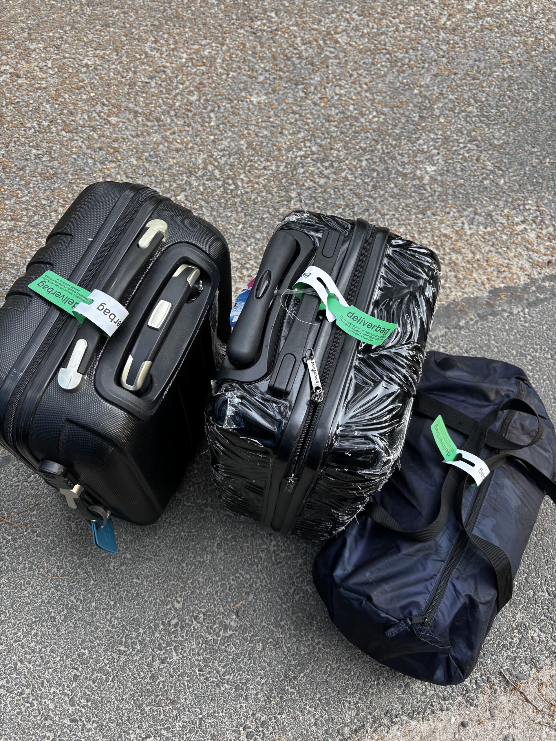 Trois bagages - une valise noire à coque rigide, une valise à coque rigide à motifs et un sac de sport foncé avec des étiquettes vertes "gearstage" - attendent sur un trottoir accidenté, prêts à être transportés vers l'Europe ou stockés en toute sécurité lors de votre prochain événement.