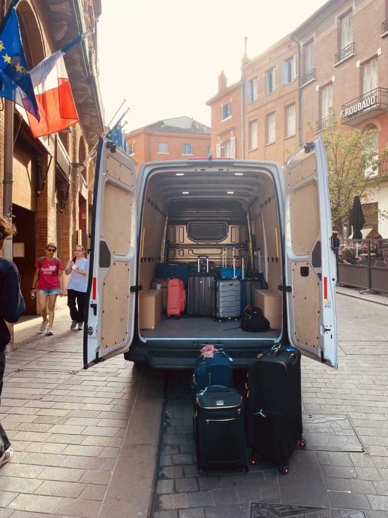 Une camionnette blanche, dont les portes arrière sont ouvertes, est garée dans une rue pavée, proposant la consigne bagages France. Plusieurs valises sont à l'intérieur et sur le sol derrière la camionnette, tandis que des gens marchent à proximité et que des drapeaux français sont accrochés à un bâtiment sur la gauche. Une camionnette blanche, dont les portes arrière sont ouvertes, est garée dans une rue pavée, proposant la consigne bagages France. Plusieurs valises sont à l'intérieur et sur le sol derrière la camionnette, tandis que des gens marchent à proximité et que des drapeaux français sont accrochés à un bâtiment sur la gauche.