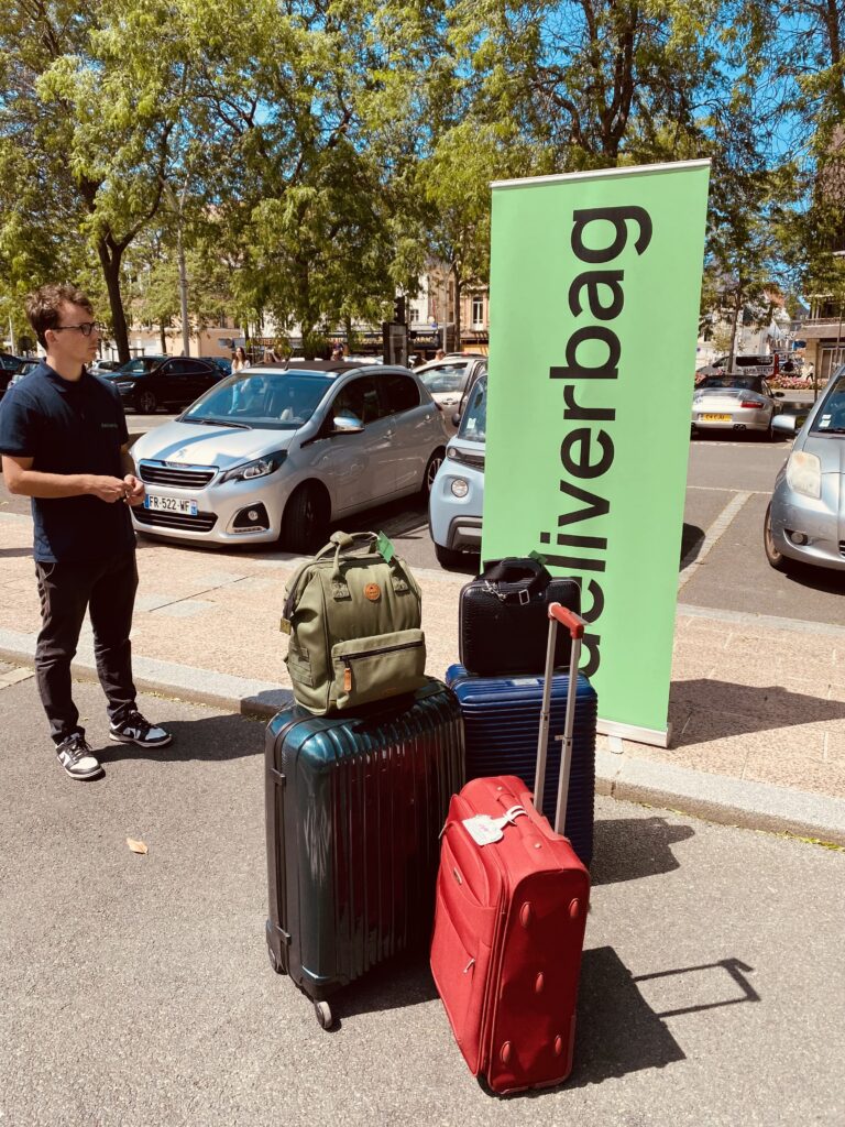 Un homme se tient près de quatre valises et d'un sac à dos dans un parking, à côté d'un panneau vert "deliverbag", indiquant livraison bagages sécurisée. Des voitures garées et des arbres sont visibles à l'arrière-plan. Un homme se tient près de quatre valises et d'un sac à dos dans un parking, à côté d'un panneau vert "deliverbag", indiquant livraison bagages sécurisée. Des voitures garées et des arbres sont visibles à l'arrière-plan.