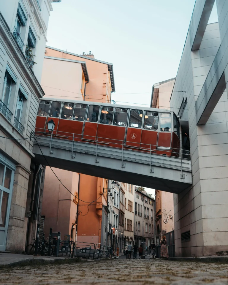 Un tramway rouge circule sur une voie surélevée entre des bâtiments dans une rue européenne étroite, avec des piétons marchant en contrebas et une architecture aux couleurs pastel. À proximité, des voyageurs utilisent la consigne bagages France pour un stockage sécurisé pendant leur voyage.