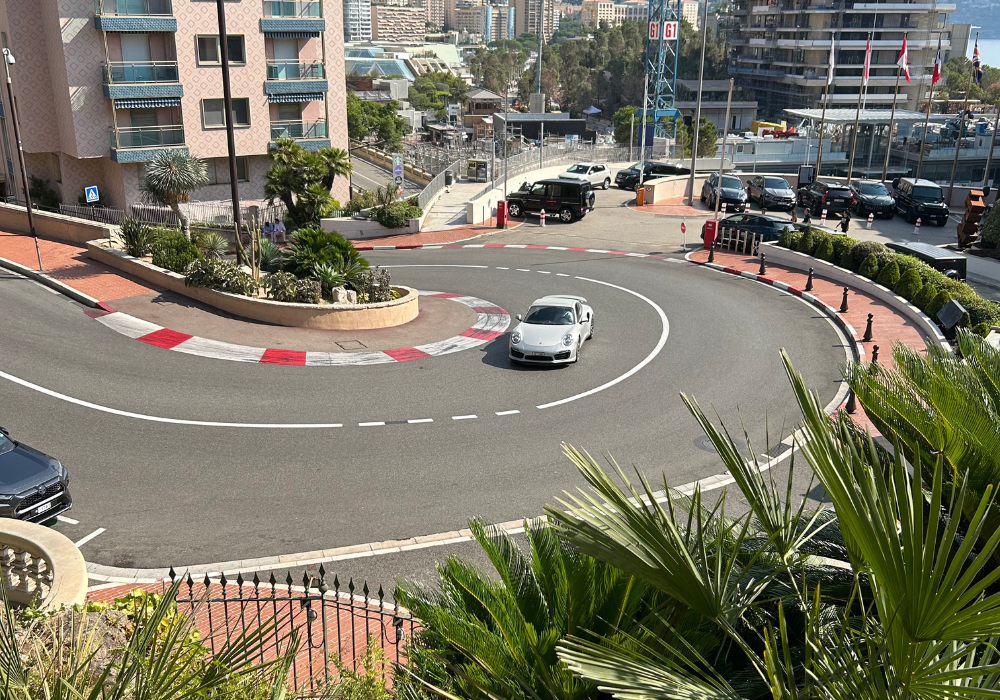 Une voiture de sport argentée négocie un virage serré en épingle à cheveux dans une rue de la ville bordée de bordures rouges et blanches, avec des palmiers et des bâtiments en arrière-plan - parfait pour les événements nécessitant un portage bagages événements ou un stockage bagages sécurisé.