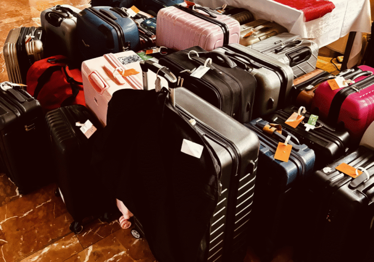 Un groupe de valises assorties de différentes couleurs et tailles, dont certaines portent une étiquette de bagage, sont empilées sur un sol en marbre à côté d'une table recouverte d'une nappe blanche et rouge, illustrant un service de bagagerie mobile ou de stockage de bagages sécurisé.