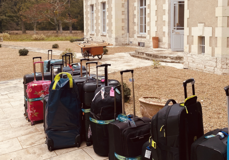 Une rangée de valises et de sacs aux poignées allongées est alignée sur un patio en pierre à l'extérieur d'un grand bâtiment, mettant en valeur notre service de bagagerie mobile, avec une brouette et des plantes en pot visibles à l'arrière-plan. Une rangée de valises et de sacs aux poignées allongées est alignée sur un patio en pierre à l'extérieur d'un grand bâtiment, mettant en valeur notre service de bagagerie mobile, avec une brouette et des plantes en pot visibles à l'arrière-plan.