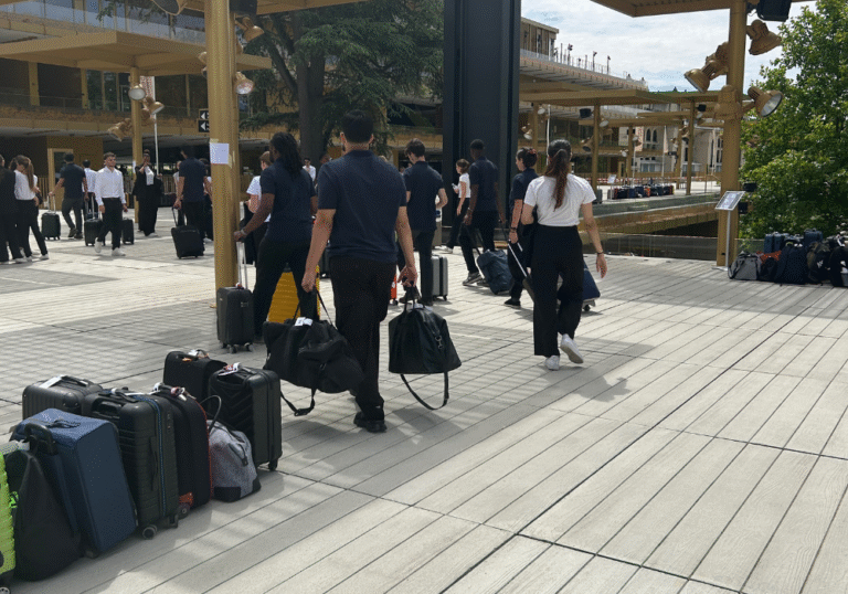 Des personnes marchent à l'extérieur sur une terrasse en bois, chacune portant ou tirant des bagages. Plusieurs valises sont regroupées sur le côté. Cette scène animée et organisée, au milieu d'arbres et de bâtiments modernes, met en valeur un service de bagagerie mobile pour des voyages sans encombre.