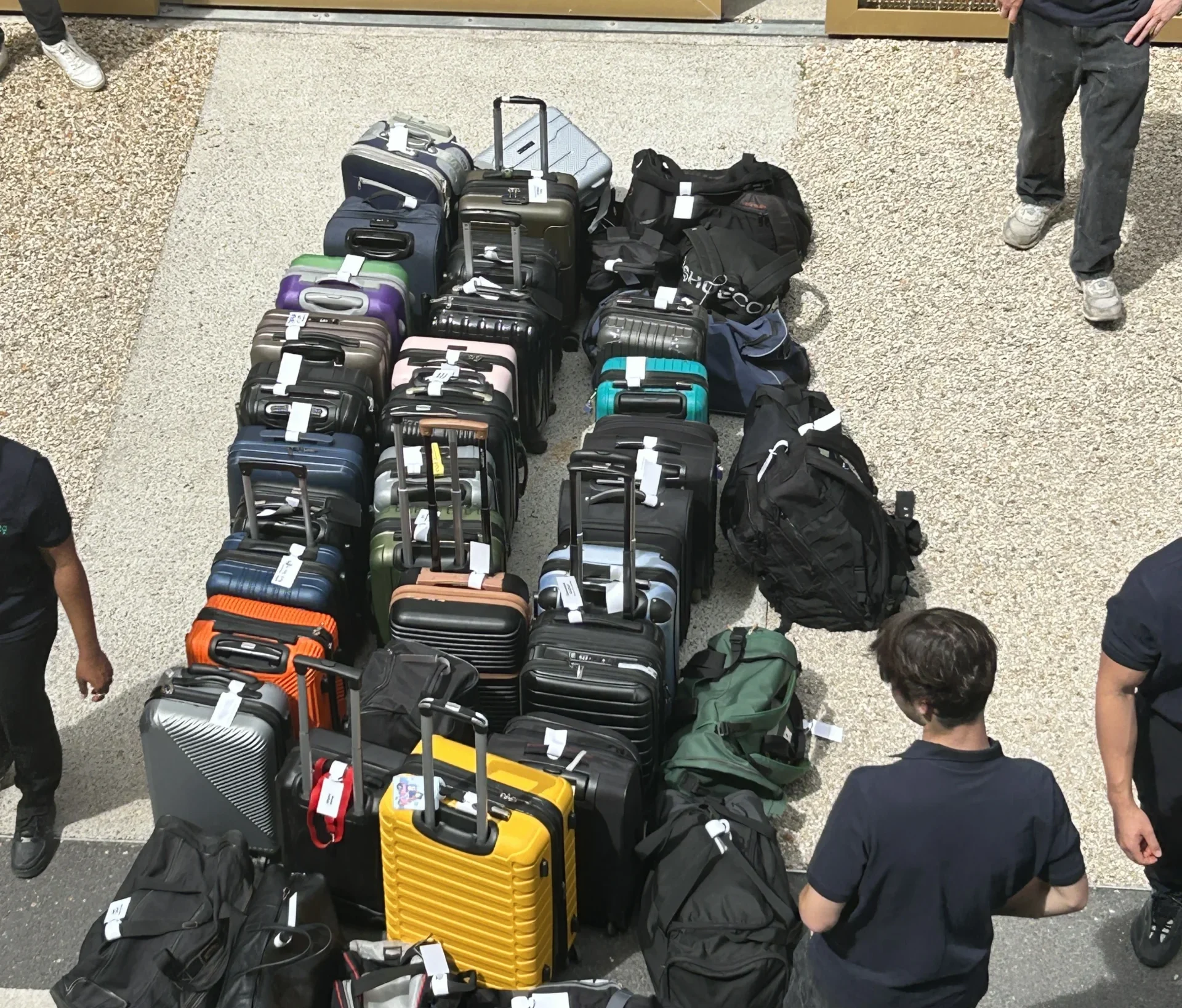 A group of people stand around neatly arranged luggage, mainly suitcases of different colours and sizes forming the letter "M" in mid-air, highlighting the luggage-carrying events on a paved and gravelled surface.