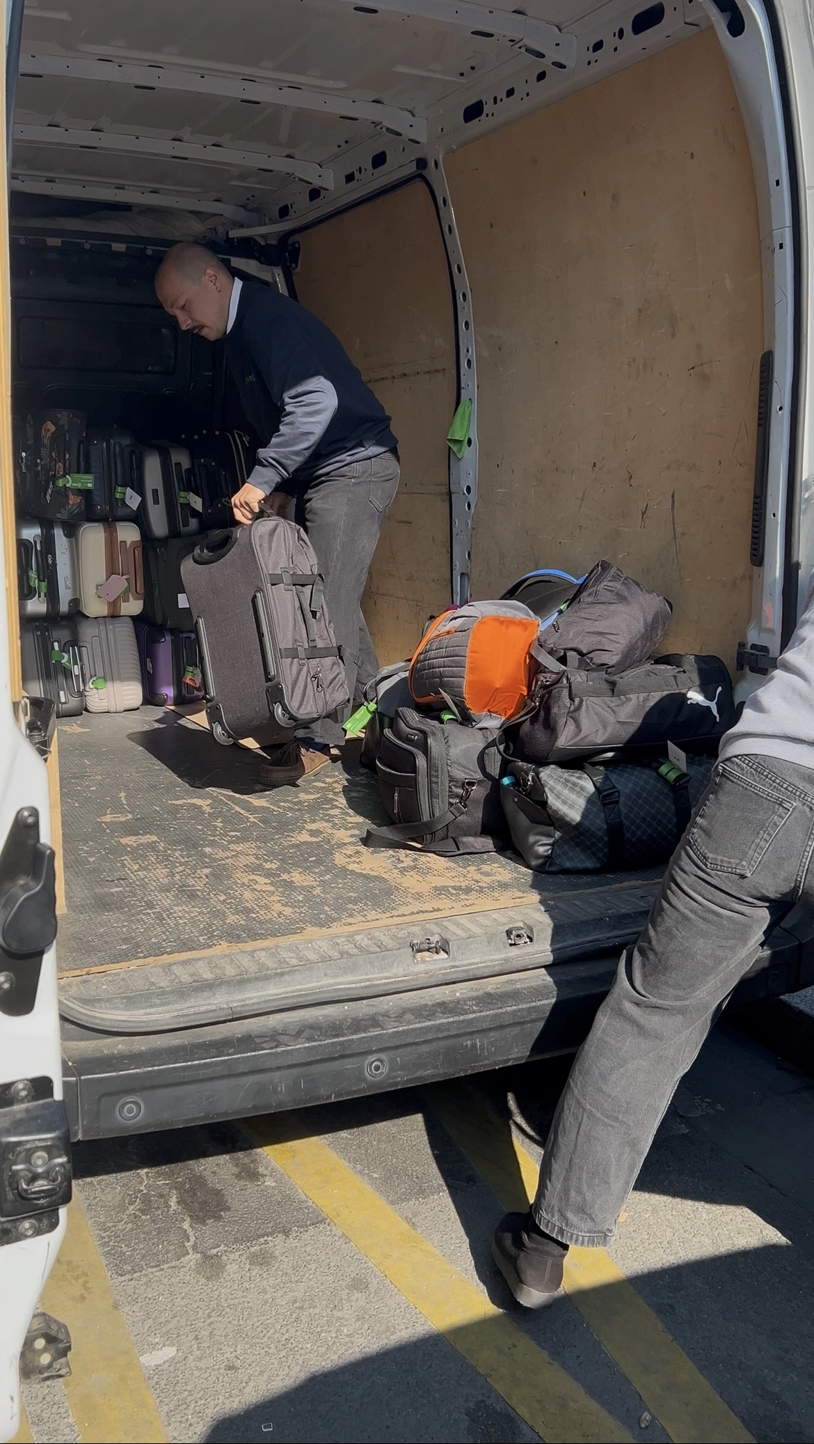Une personne charge des valises noires et des sacs de sport à l'arrière d'une camionnette blanche, tandis qu'une autre se tient à proximité. Les portes ouvertes de la camionnette révèlent d'autres bagages, mettant en évidence la consigne bagages France sous la lumière du soleil.