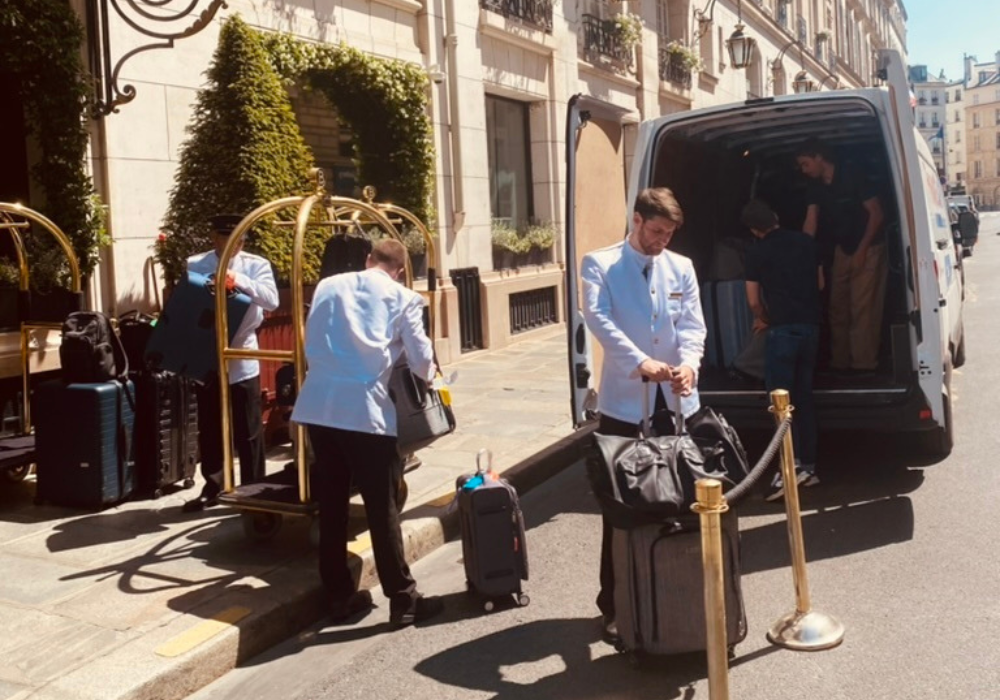 Des employés en uniforme blanc chargent des bagages sur un chariot et dans une camionnette blanche garée à l'extérieur d'un bâtiment dans une rue ensoleillée, offrant un service de bagagerie mobile. Deux personnes placent les sacs dans la camionnette tandis qu'une autre range les valises sur le trottoir.