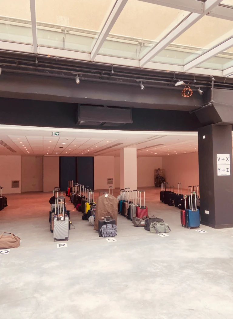 Rows of suitcases and bags with vertical handles are lined up on a concrete floor in a spacious, well-lit interior. The organisation of the storage reflects the efficiency of the event luggage, with rows identified by large letters for easy identification.