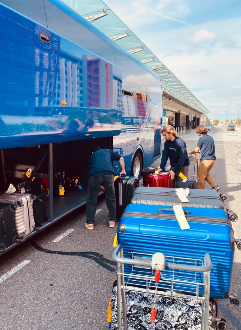 Three people load suitcases and event bags into the storage compartment of a blue coach at a terminal. Several bags, stacked on a trolley nearby, demonstrate the efficiency of the mobile luggage service. In the background, buildings and a clear sky.