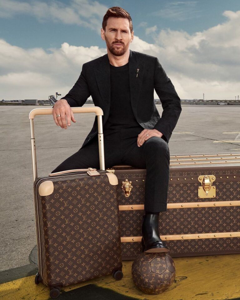 Un homme en costume noir est assis sur une grande malle Louis Vuitton, une main sur une valise assortie - parfaite pour transporter des bagages en Europe - et un pied posé sur un ballon de football Louis Vuitton, sur fond de trottoir vide et de ciel nuageux.