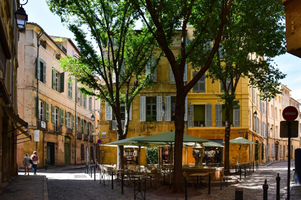 Une rue européenne ensoleillée avec de grands arbres, des tables de café en plein air sous des parapluies verts et des bâtiments historiques jaunes aux fenêtres fermées. Deux personnes marchent sur la route pavée à l'arrière-plan.