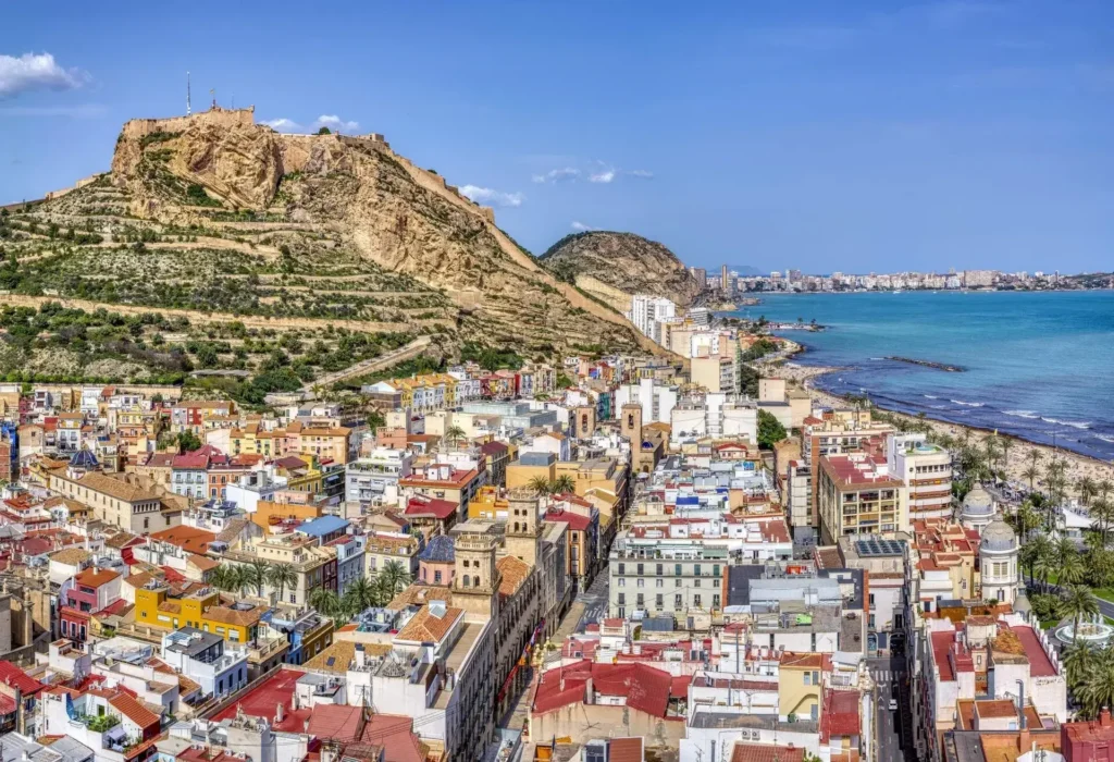 Bâtiments colorés et paysage urbain d'Alicante, Espagne, avec le château de Santa Bárbara sur une colline rocheuse surplombant la ville et la mer Méditerranée sous un ciel bleu clair.