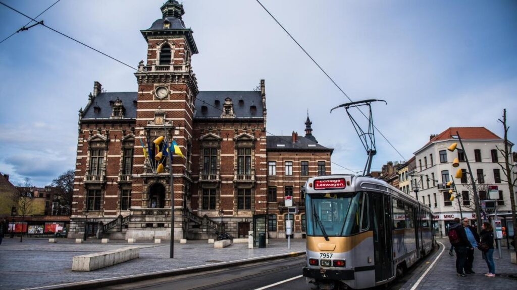 Un tramway moderne passe devant un bâtiment historique en briques avec une tour d'horloge sur une place de la ville, avec des drapeaux belges et des câbles de tramway visibles dans un ciel nuageux.