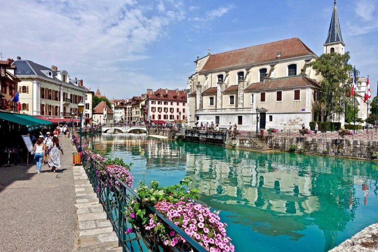 Un canal pittoresque à l'eau turquoise traverse une ville européenne bordée de bâtiments colorés, de fleurs, de piétons et d'une église au clocher élevé sous un ciel partiellement nuageux.