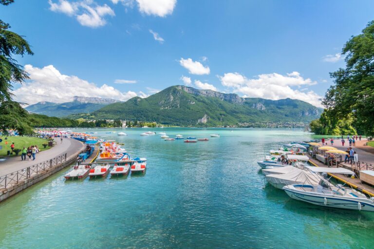 Vue panoramique d'un lac turquoise avec des bateaux amarrés le long des deux rives, des personnes marchant sur des sentiers et des montagnes verdoyantes en arrière-plan, sous un ciel partiellement nuageux.