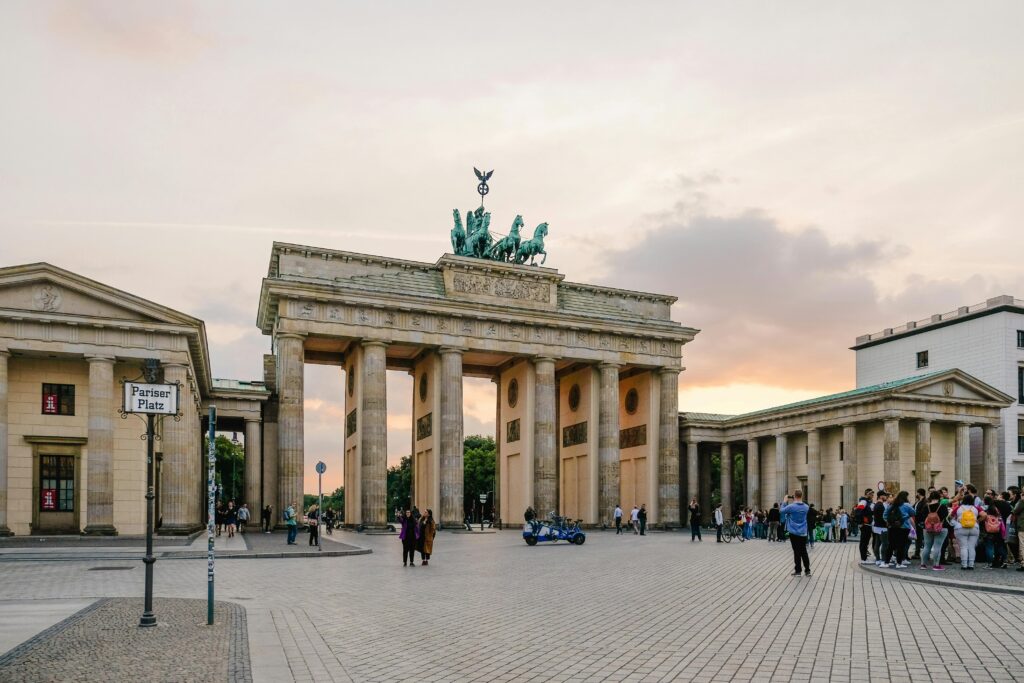 La porte de Brandebourg à Berlin au coucher du soleil, avec des groupes de personnes rassemblées autour et un ciel aux teintes orange et violettes. Le monument historique se dresse au centre d'une grande place ouverte.