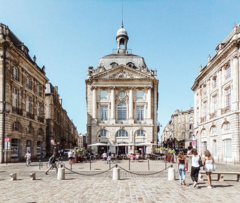 Un grand bâtiment historique avec une horloge et un dôme se dresse sur une place pavée ensoleillée, entourée d'architectures classiques similaires. Les gens marchent, font du vélo et s'assoient à des tables en plein air devant le bâtiment.
