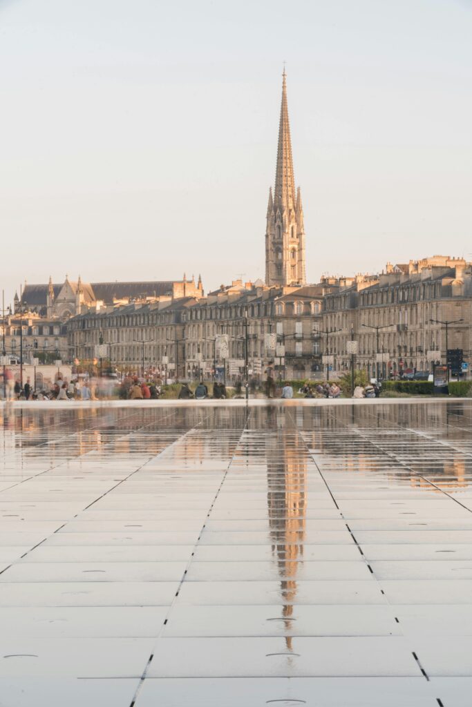 Une haute église à la flèche pointue se reflète dans une grande place carrelée et humide, avec des bâtiments historiques et des personnes à l'arrière-plan sous une douce lumière du jour.