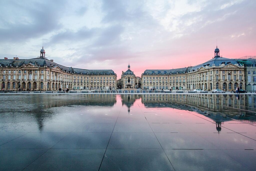 Bâtiments historiques symétriques se reflétant dans un grand miroir d'eau peu profond au coucher du soleil, avec un ciel rose et bleu au-dessus de la tête. La scène est paisible et met en valeur l'architecture européenne classique.