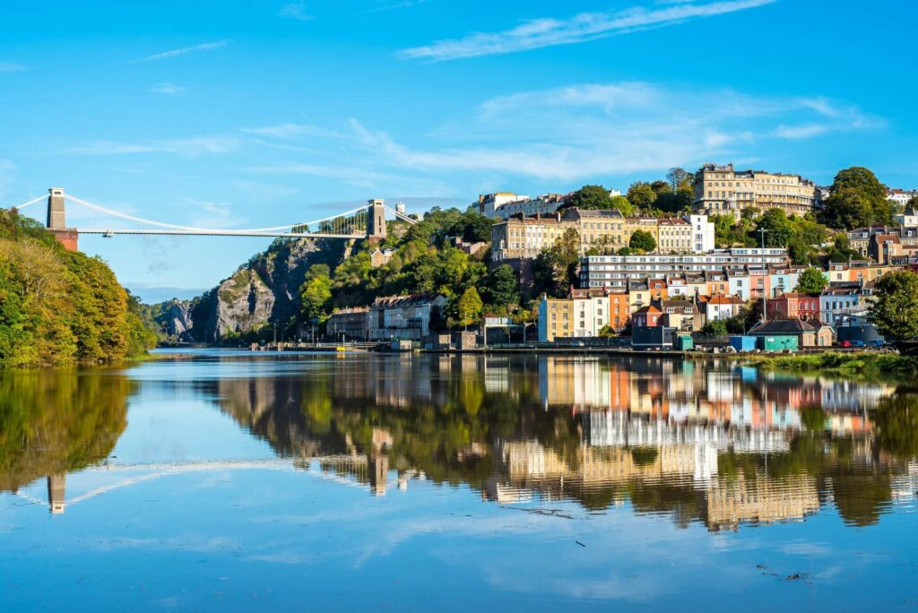 Vue du pont suspendu de Clifton au-dessus des gorges de l'Avon à Bristol, en Angleterre, avec des maisons colorées et de la verdure se reflétant dans la rivière calme sous un ciel bleu vif.