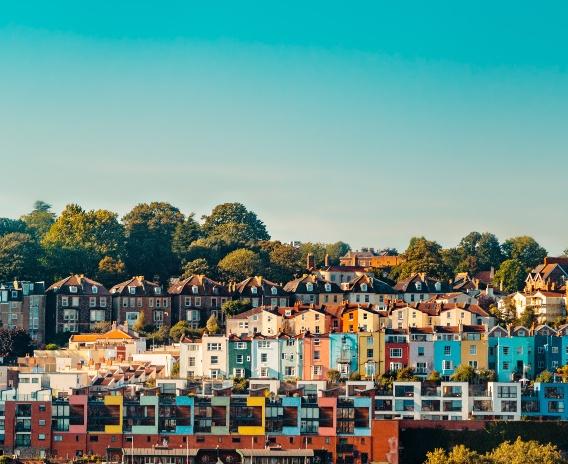 Des maisons et des immeubles colorés se dressent en rangs serrés sur le flanc d'une colline, entourés d'arbres verts sous un ciel bleu limpide.