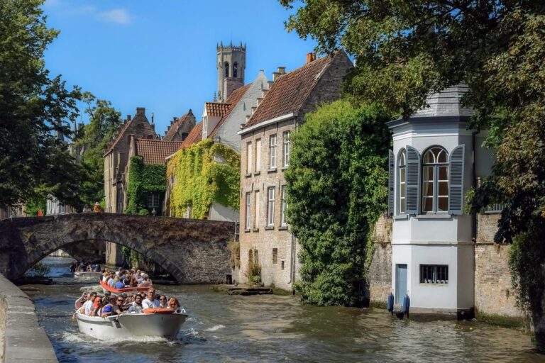 Un bateau rempli de touristes descend un canal bordé de bâtiments historiques en briques et de feuillages verts à Bruges, en Belgique. Un pont de pierre enjambe l'eau et un clocher s'élève à l'arrière-plan sous un ciel bleu.