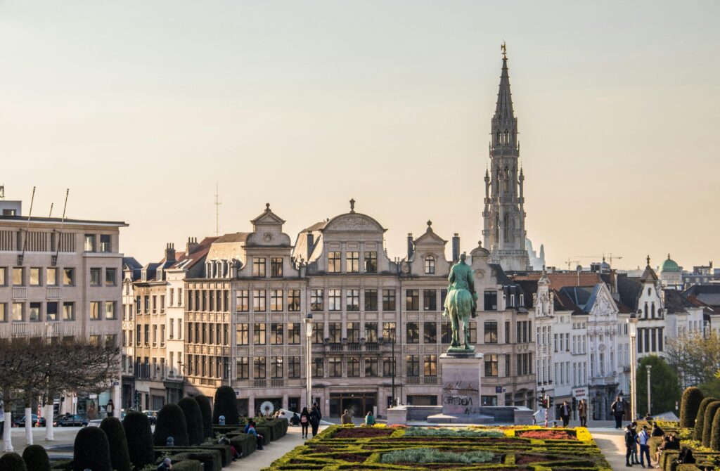 Paysage urbain de Bruxelles vu d'un jardin, avec des bâtiments historiques, la statue d'un cavalier à cheval et la haute flèche de l'hôtel de ville de Bruxelles à l'arrière-plan. Des personnes se promènent et se détendent dans le parc.