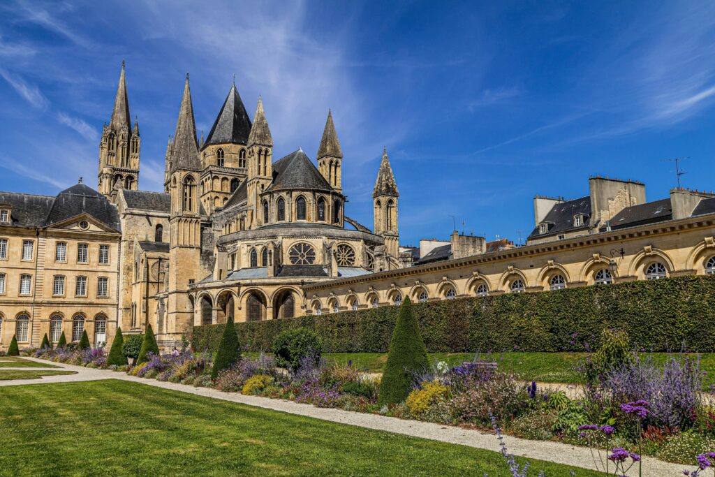 A large historic stone abbey, with tall spires and ornate towers, stands behind a manicured garden, blooming with purple and green plants, under a bright blue sky with billowing clouds.