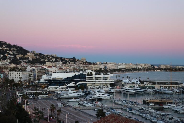 Un port de plaisance rempli de yachts et de voiliers se trouve à côté d'une ville côtière au coucher du soleil. Des bâtiments bordent le rivage et des collines s'élèvent à l'arrière-plan sous un ciel rose et bleu.