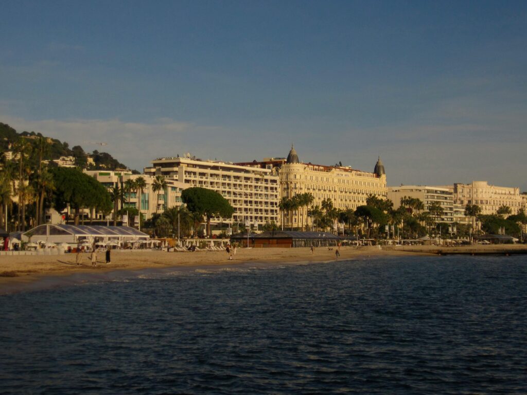 Vue d'une plage de sable à Cannes, France, avec des gens marchant le long du rivage, des palmiers et d'élégants bâtiments historiques bordant le front de mer, sous un ciel bleu clair. Vue d'une plage de sable à Cannes, France, avec des gens marchant le long du rivage, des palmiers et d'élégants bâtiments historiques bordant le front de mer, sous un ciel bleu clair.