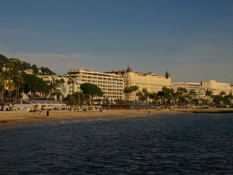 Vue d'une plage de sable à Cannes, France, avec des gens marchant le long du rivage, des palmiers et d'élégants bâtiments historiques bordant le front de mer, sous un ciel bleu clair.