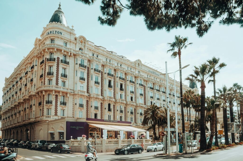Vue de la rue du grand hôtel Carlton à Cannes, France, avec une architecture ornée, des palmiers, des voitures garées, des scooters, et des personnes dînant dans un café sur le trottoir sous un auvent violet par une journée ensoleillée.