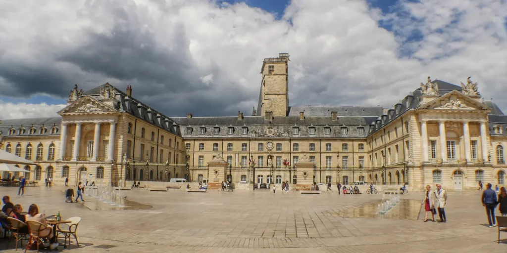 Un grand bâtiment historique avec une tour centrale et des ailes symétriques, situé autour d'une vaste place pavée. Les gens se promènent et s'assoient à des tables en plein air sous un ciel partiellement nuageux.