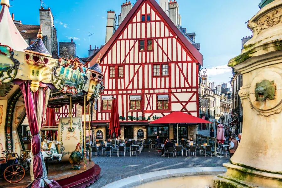 Un bâtiment à colombages coloré avec des rayures rouges et blanches se dresse derrière un café en plein air, à côté d'un manège et d'une fontaine en pierre sur une place européenne animée sous un ciel bleu.
