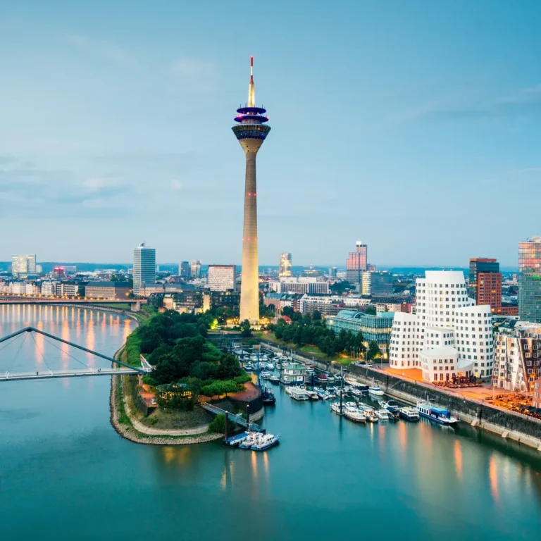 Vue de la ville de Düsseldorf, en Allemagne, avec le Rhin, la tour de télévision Rheinturm, des bâtiments modernes et des bateaux amarrés au bord de l'eau au crépuscule.