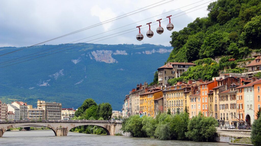 Une rangée de bâtiments colorés borde une rivière, traversée par un pont de pierre. Quatre téléphériques sphériques glissent au-dessus, entourés de collines verdoyantes et de montagnes escarpées sous un ciel partiellement nuageux.